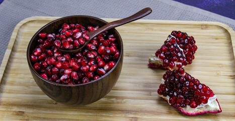Red pomegranate seeds close up in a wooden bowl with a wooden spoon. Two pieces of pomegranate with ripe red seeds on a wooden tray.