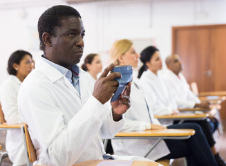 Interested African American in white coat taking photos or filming at smartphone at lecture during medical conference