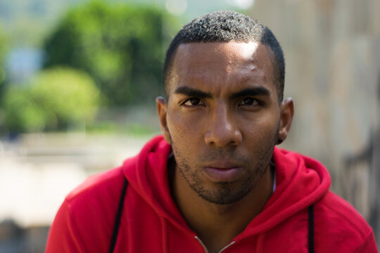 Hombre acuerpado de piel morena en un parque mirando serio al frente con un buso rojo y una mochila sudando mientras el sol cae sobre &eacute;l generando sombras en su cuerpo