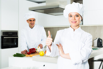 Young woman professional cook in uniform showing thumb up on kitchen