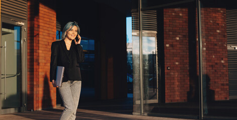 Fototapeta premium Blue haired businesswoman walking in the street and discuss on phone holding a laptop and smile