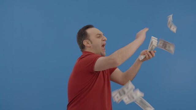 Young man with beard in front of a blue background who is cheerfully gesturing while handing out money. Concept of to pour money without thinking. It makes it rain by throwing money into the air.