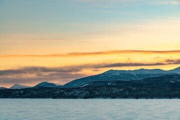 A stunning winter view of a frozen lake in northern Canada with bright orange, yellow pastel cloudy sky in the background.