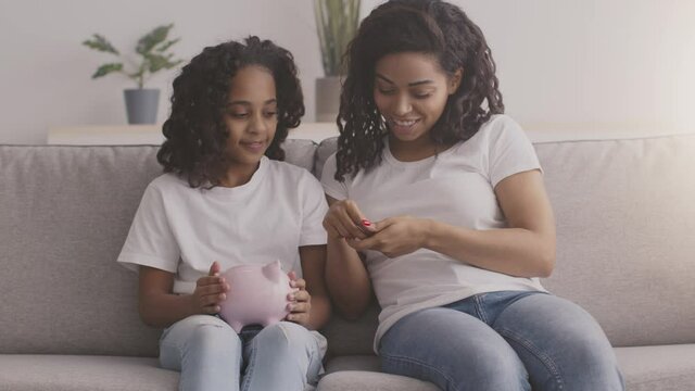 Young black mother putting money into daughters piggybank and talking about financial literacy at home, slow motion