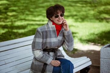 Young cheerful woman in coat talking on the phone while sitting on a bench in spring