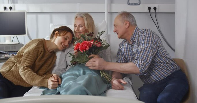 Happy senior husband and young daughter visiting and cheering woman lying in bed at hospital ward