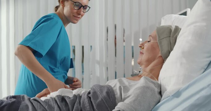 Female nurse supporting and holding hand of sick woman having cancer and lying in bed at hospital