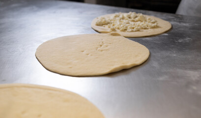 Dough circles for dumplings and pies are rolled out on a metal board, one of the circles is filled with cheese. Raw and fresh baked goods.