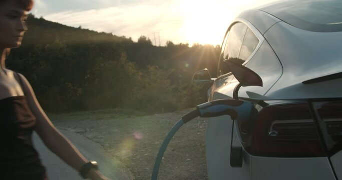 Girl plugs out the charging cable from her electric car during sunset. Handheld video