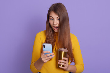 Fototapeta premium Photo of surprised woman being surprised, shocked to see new post, connected to wireless internet, reads astonishing news, drinks coffee or tea from thermo mug, wearing yellow jumper.