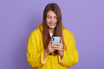 Portrait of happy Caucasian woman feeling happiness and typing smart phone isolated on lilac background, cute girl wearing casual yellow jacket, connects internet, surfing.