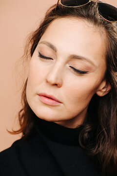Tired middle aged woman resting with her eyes closed. She has subtle cat eye makeup, she wears black shirt. Over peach background.