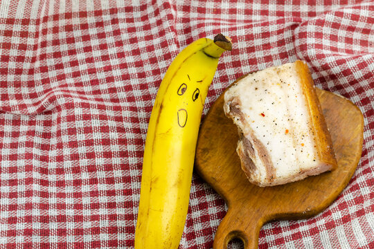 Banana with a smiley face and a piece of salty fat on a white and red checkered cloth.