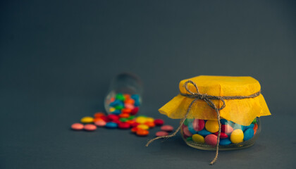 Chocolate candies, small multi-colored in a jar with a bow, on a gray background and with a yellow lid.