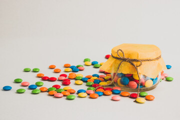 Chocolate candies, small multi-colored in a jar with a bow, on a gray background and with a yellow lid.