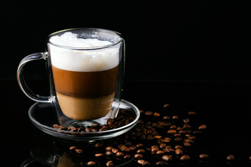 Cappuccino coffee in a transparent cup on a black background and scattered coffee beans.