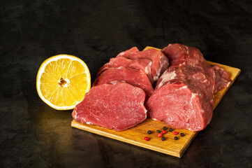 Fresh raw veal tenderloin meat in large chunks and sliced medallions on a wooden chopping board on a dark background.