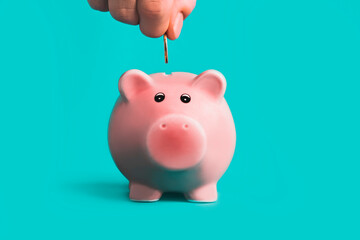 Hand putting coin into the pink piggy bank. Turquoise background. Saving money concept, business finance and investment.