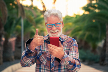 Smiling senior man using technology with smartphone for video call with family. Standing outdoor in a public park at sunset. Happiness in his face