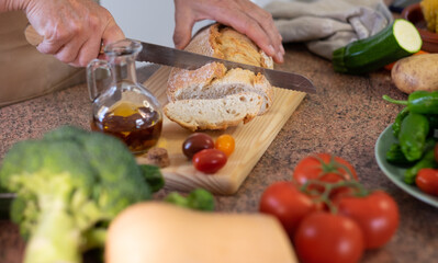 Slices of rustic bread on the wooden cutting board and two woman's hands preparing a snack with a tomato bruschetta and spicy oil. Assortment of fresh vegetables on the table