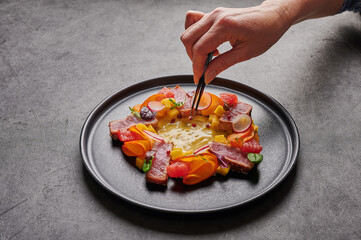 Woman's hand lays out the grain of red pepper with tweezers to homemade salad with tuna steak, mango sauce, herbs and spices on a dark plate, close up, food decorating concept