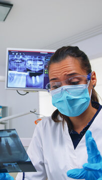 Patient pov in dental office explaining treatment of teeth cavity, dentist pointing on x-ray image. Stomatology doctor wearing protective mask and gloves, working in modern stomatological clinic