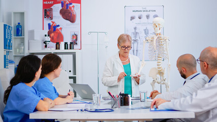 Elderly woman doctor showing the work of human's hand on skeleton anatomical model. Specialist medic explaining diagnosis to colleagues standing in front of desk in hospital meeting room.