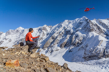 A beautiful landscape with mountains, a huge blue glacier and a middle-aged mountaineer sitting with a walkie-talkie in his hands calls the rescue service waiting for an ambulance helicopter