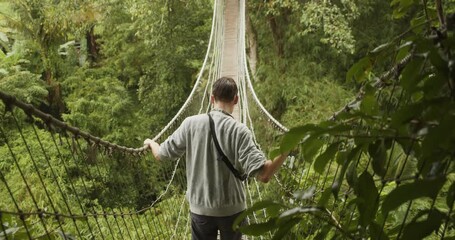 Back close up view of a man making his way across an unstable rope jungle bridge in the rain Young man crossing a hanging bridge in Bali, Indonesia