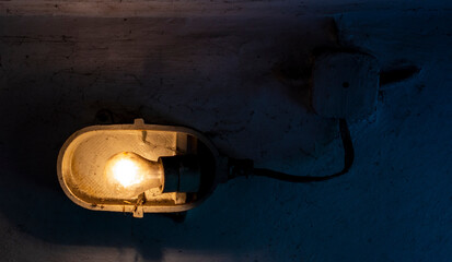 Industrial old ceiling lamp (storage or factory lamp) with vintage switched on bulb on dirty wall in a dark warehouse, basement or attic

