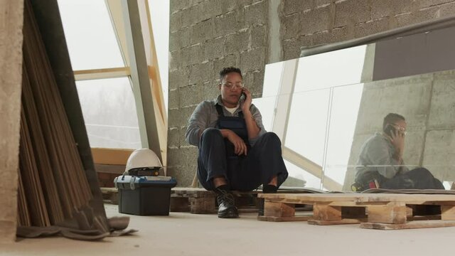 Long tracking right of Mixed-Race female construction worker sitting on pallet in room under renovation, talking on smartphone, having break