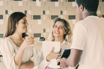 Three cheerful designers standing together and discussing work. Serious African American man showing something on tablet screen to smiling Caucasian women. Communication and teamwork concept