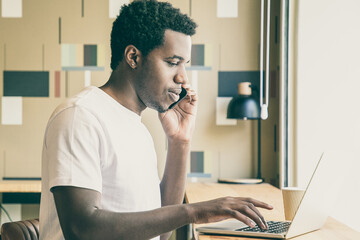 Focused young entrepreneur working at laptop and talking on mobile phone in co-working space. African American guy sitting at desk with computer and coffee. Medium shot. Communication concept