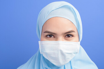 Young muslim woman with hijab wearing a surgical mask over blue background studio.