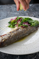 whole grilled fish on a plate with salad on a dark background vertically poured with lemon juice
