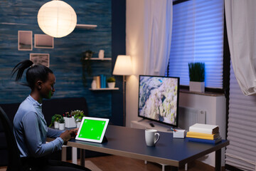 African businesswoman looking at tablet pc in the course of working from home late at night in home offce sitting at desk. Using mockup chroma key display computer.