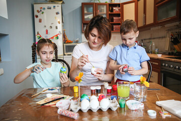 Mom with two children decorate Easter eggs sitting at the table at home in the kitchen