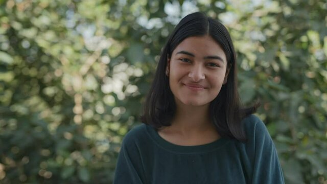 close view shot of a cheerful young asian Indian gorgeous looking female with smile on the face staring at lens in a broad daylight, trees in background