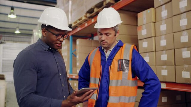 Smiling caucasian male warehouse worker and african manager looking at digital tablet discussing in storage factory
