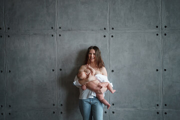 A young mother with a thoughtful look in white, down to the shoulders, shirt and jeans holds the baby and breastfeeds.