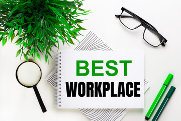 On a white background lies a notebook with the word BEST WORKPLACE, glasses, a magnifying glass, green markers and a green plant