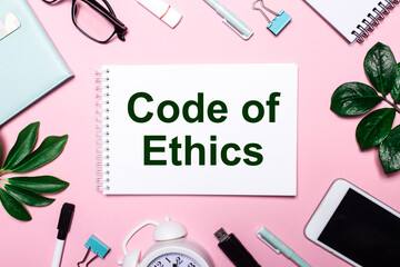 CODE OF ETHICS is written in a white notebook on a pink background surrounded by business...