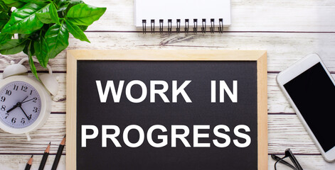 WORK IN PROGRESS written on a black background near pencils, a smartphone, a white notepad and a...