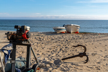 small motor winch and anchors and small wooden fishing rowboats on a sandy beach