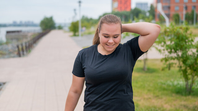 An obese young woman jogging outdoors. Fat beautiful smiling girl in a black tracksuit is engaged in fitness for weight loss on the waterfront. A woman runs on a summer day.