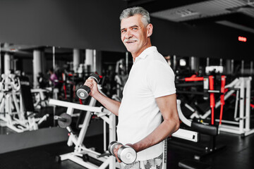 Fototapeta na wymiar Portrait of an old grandfather in good physical shape, he trains in the gym with dumbbells in his hands and smiles at the camera