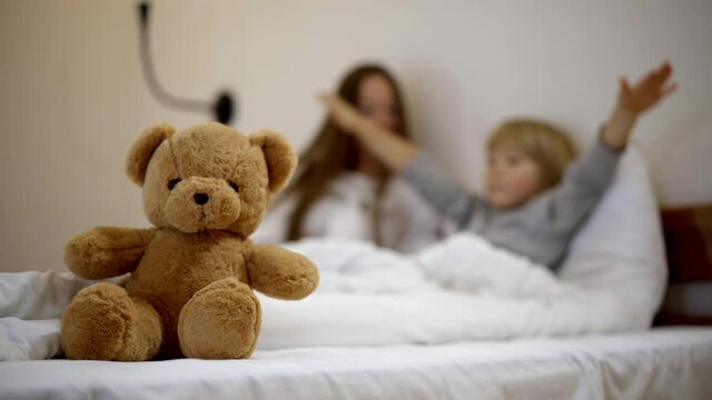 Close-up of teddy bear with blurred mother and son lying in bed talking and gesturing. Happy Caucasian woman and boy sharing at bedtime at home. Unity and bonding concept.