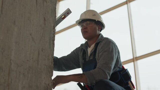 Low angle medium long of Mixed-Race female builder wearing protective goggles and helmet, using bubble level tool on concrete pillar in construction in progress