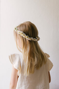 Blonde haired girl wearing a daisy crown seen in front of a empty neutral white background wearing a white summer blouse with her back turned to the camera.