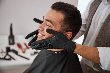 Handsome male resting in the barber shop while barber putting cosmetic oil on skin face